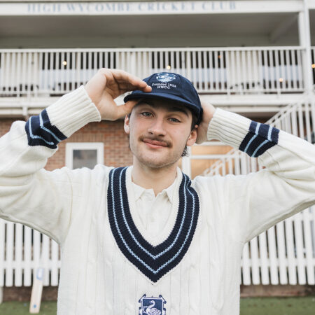 This image shows a cricket playing putting his baggy cap on whilst wearing a white cable-knit, wool traditional jumper with blue v-neck trim.